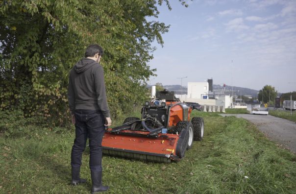 Ein Mann auf einer Wieser neben einer Straße, vor ihm steht ein roter Mähroboter