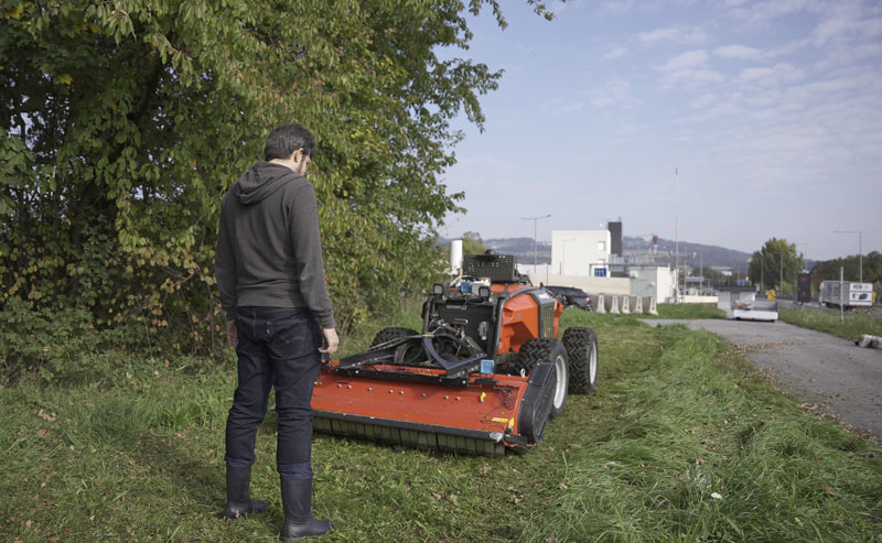 Ein Mann auf einer Wieser neben einer Straße, vor ihm steht ein roter Mähroboter